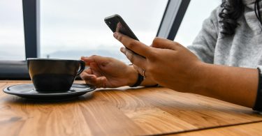 La mano de un adulto sostiene un teléfono móvil mientras lee algo en la pantalla, y con la otra busca levantar la taza de cafe.