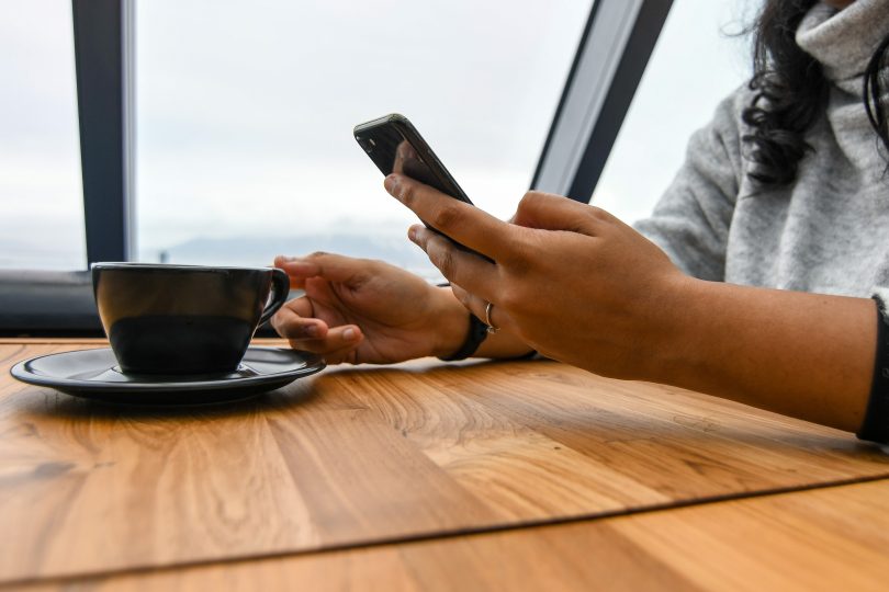 La mano de un adulto sostiene un teléfono móvil mientras lee algo en la pantalla, y con la otra busca levantar la taza de cafe.
